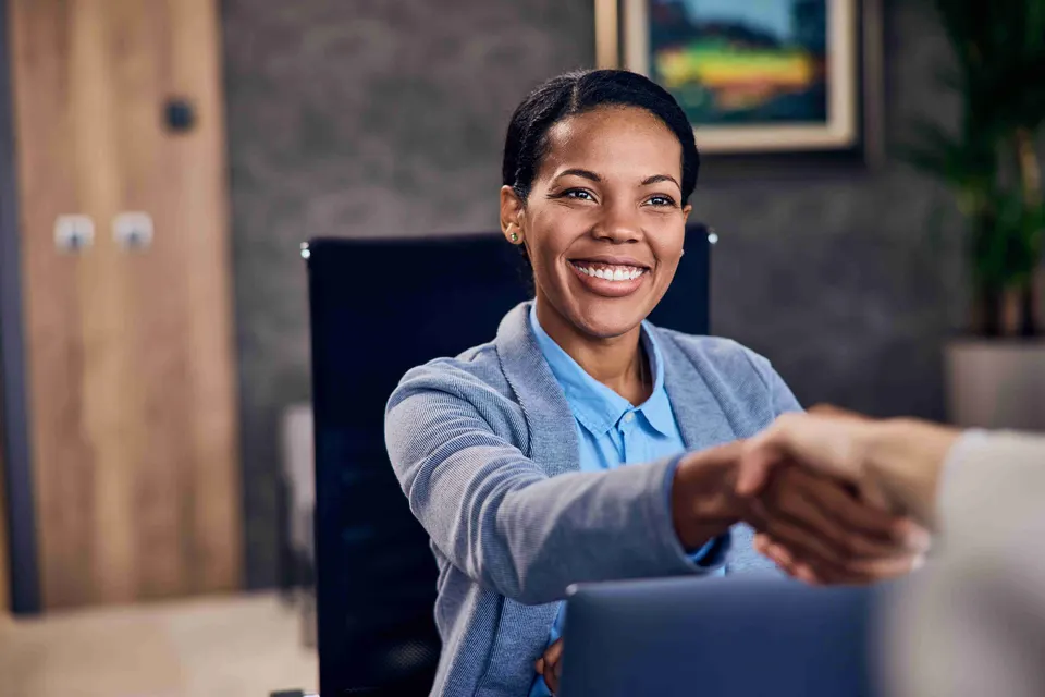 Smiling woman in a blue blazer shaking hands across a desk